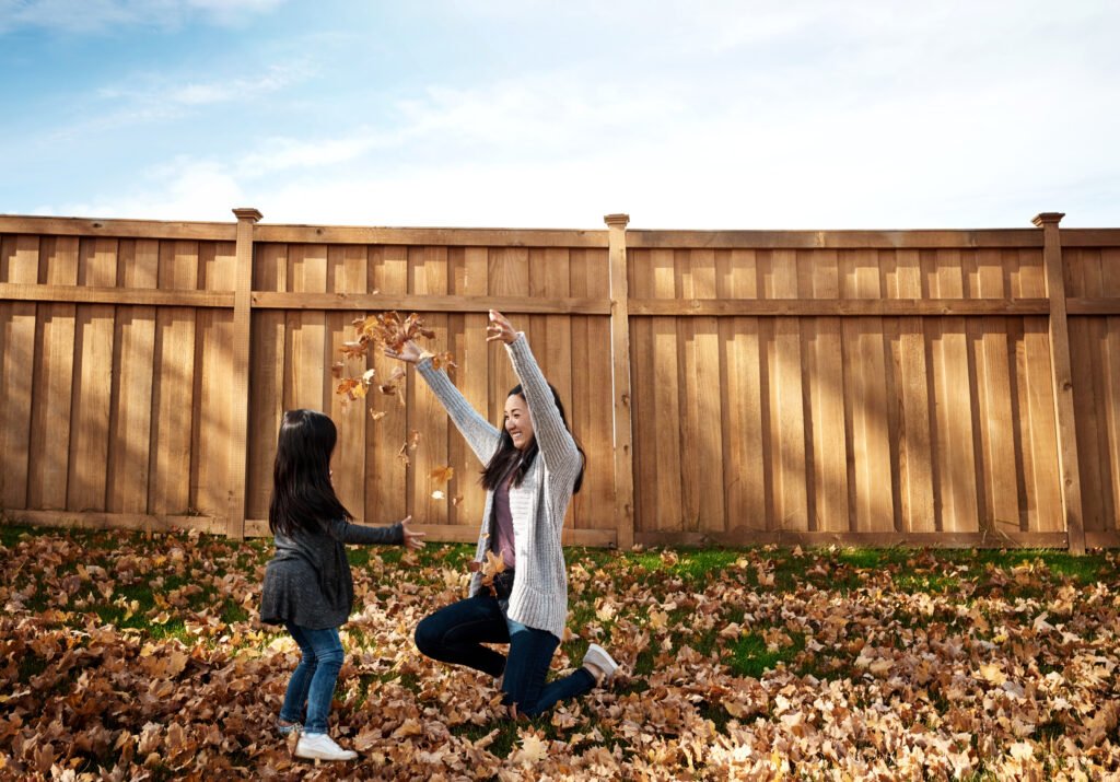 Mother and child playing in autumn leaves in front of a wood privacy fence